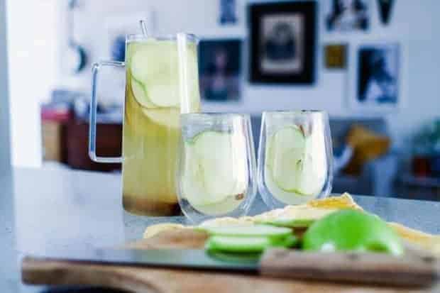 Pitcher and 2 glasses of white sangria with apple slices on the counter and a cutting board with apples. Pictures hanging on the wall in the background.