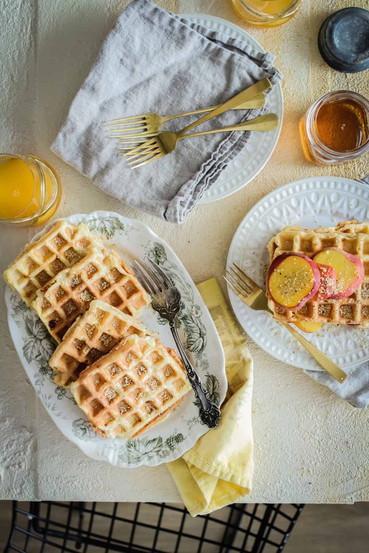 waffles on serving plate and in a stack with sliced peaches on top. Maple syrup and orange juice on the side.