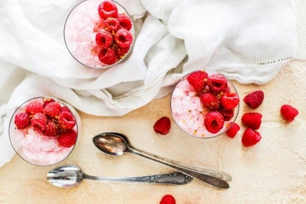 3 pink granita parfaits with fresh raspberries on top of a cream colored table with 2 metal spoons and a white linen napkin