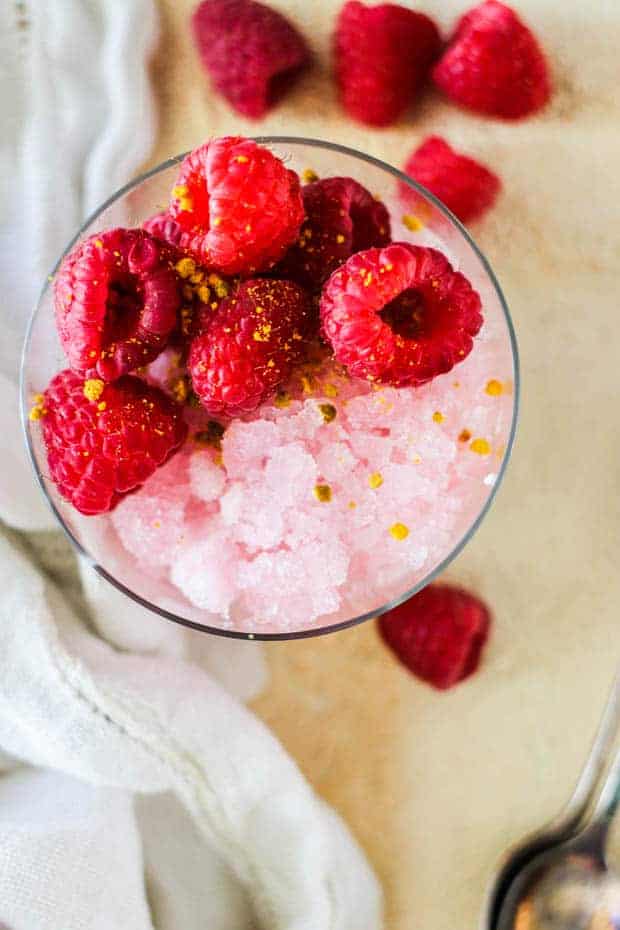 Looking down onto a table top with a glass that has pink shaved ice, fresh raspberries, ad a sprinkle of bee pollen. There are fresh raspberries, a white linen napkin, and a spoon on the table. 