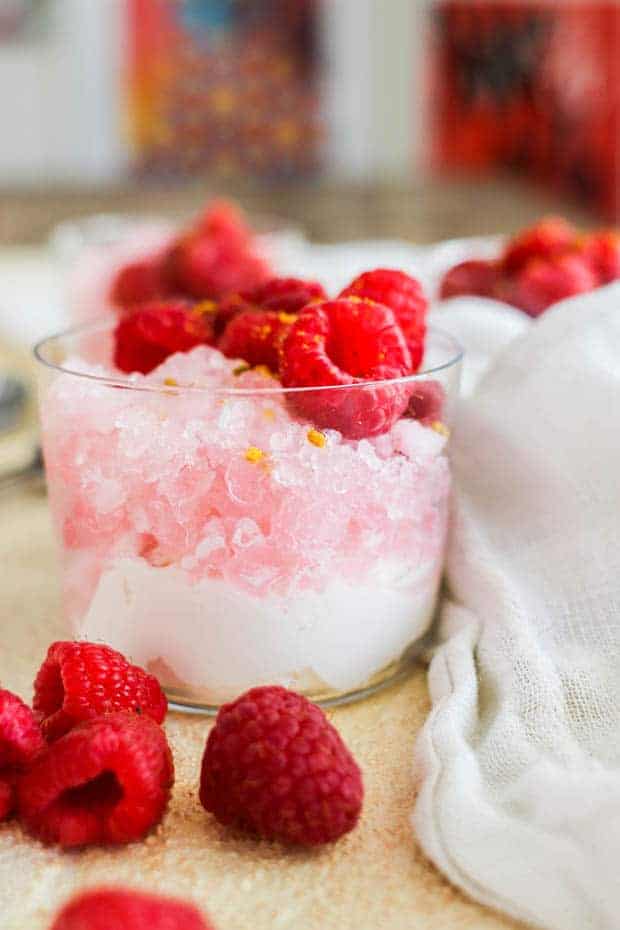A clear glass up with a layer of whipped cream, topped with pink shaved ice, then topped with raspberries on tp of a table with a white linen napkin and fresh raspberries