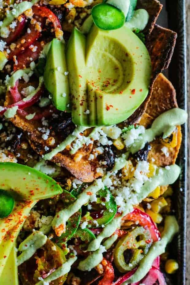 Sheet pan covered in sweet potato tortilla chips and blue corn tortilla chips then topped with fire roasted corn kernels, black beans, jalapeños, pickled red onions, thinly sliced avocado, Aleppo pepper flakes and a green roasted poblano cashew cream drizzle.