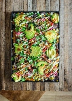 A sheet pan of nachos covered in a variety of toppings like corn, black beans, pickled onions, and avocado slices on top of a wood table top . You can also see a wooden chair in the images as well.