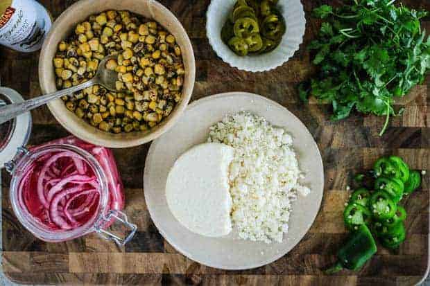 a cutting board with cilantro, sliced jalapeño peppers, a plate of crumbled Cotoja, a jar of pickled red onions, a bowl of fire roasted corn kernels, a small dish of pickled jalapeño slices, and a bottle of Pompeian Extra Virgin Olive Oil Spray