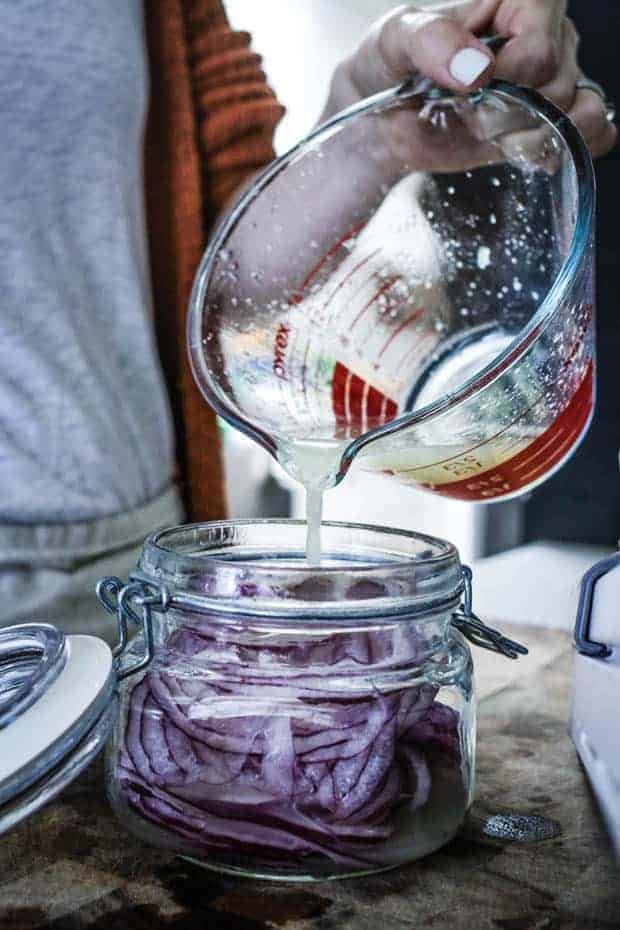 woman in white shirt and orange sweater pouring lime juice from a measuring glass into a jar of thinly sliced red onions that is setting on top of a wooden cutting board.