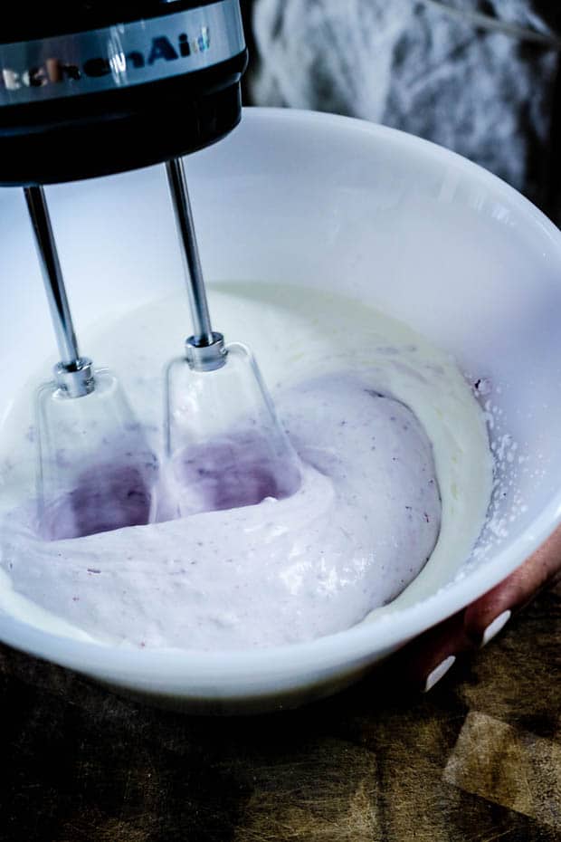 A white mixing bowl with a hand held mixer beating heavy cream. Teh whipped cream has turned half white/half purple from the addition of blueberry jam in the whipped cream 