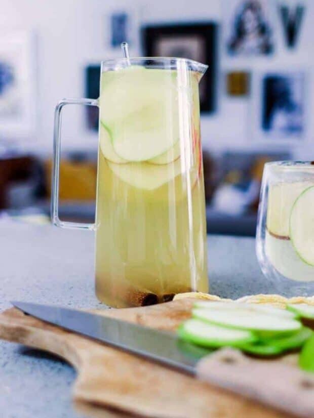 Pitcher of white sangria with apple slices on the counter and a cutting board with apples and a knife in the foreground. Artwork hanging walls in the background.