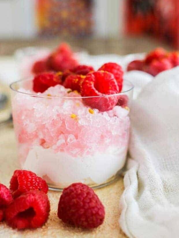 A clear glass up with a layer of whipped cream, topped with pink shaved ice, then topped with raspberries on tp of a table with a white linen napkin and fresh raspberries
