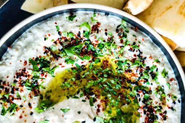 A black bowl with a white rim filled with creamy Baba Ganoush, a white dip topped with minced herbs, red pepper flakes, black sesame seeds, and a drizzle of olive oil. On the side are triangles of naan bread