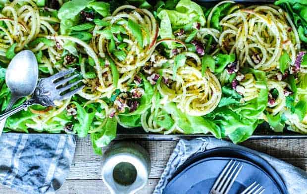 a salad made up of green lettuce, thinly sliced celery, spiralized apples tossed in poppyseed vinaigrette, and toasted walnuts all served in a rectangular metal tray with a serving fork and spoon. The tray is on top of a tobacco lathe table and also has a stack of black plates and forks, linen napkins, and a very small white stoneware pitcher filled with dressing