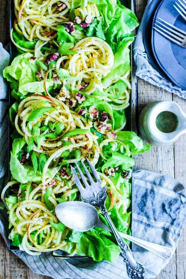 a salad made up of green lettuce, thinly sliced celery, spiralized apples tossed in poppyseed vinaigrette, and toasted walnuts all served in a rectangular metal tray. The tray is on top of a tobacco lathe table and also has a stack of black plates and forks, linen napkins, and a very small white stoneware pitcher filled with dressing