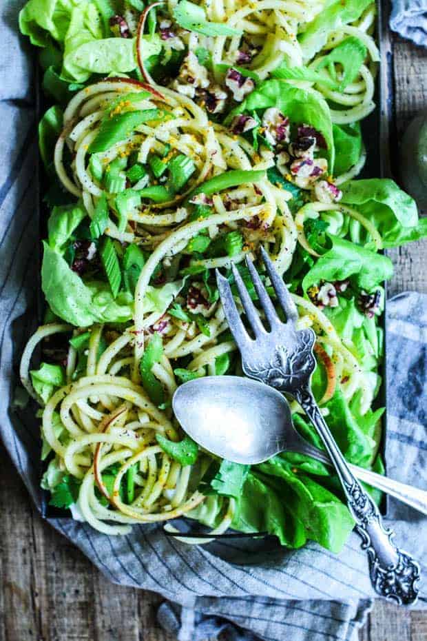 A rectangular, metal tray filled with salad greens, spiralized apples tossed in a poppyseed vinaigrette, thinly sliced celery, chopped toasted walnuts. The tray is on top of a striped black and tan linen napkin and has a serving fork and spoon laid across each other on the top. Everything is sitting on top of a wood tobacco lathe table. 