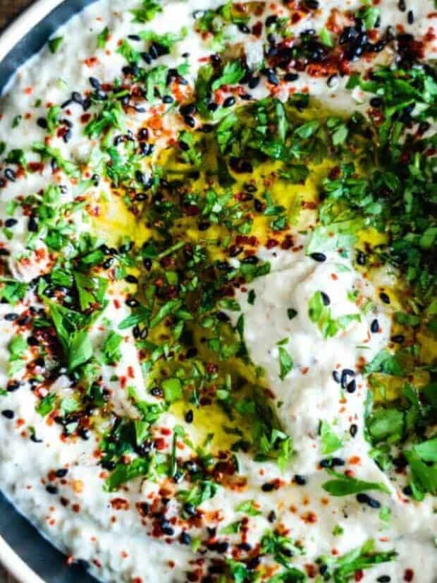 A black bowl with a white rim filled with creamy Baba Ganoush, a white dip topped with minced herbs, red pepper flakes, black sesame seeds, and a drizzle of olive oil