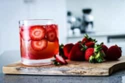 A red drink in a clear glass with ice cubes and slices of strawberries floating in it. Teh glass is on top of a wooden board that is placed on a grey kitchen counter and there are sliced berries next to the drink