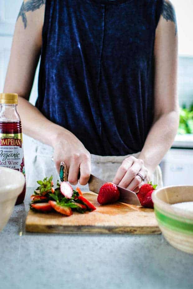 A woman in a blue tank top slicing strawberries son a wooden cutting board. There is a cream bowl with a green rim and a bottle of Pompeian red Wine Vinegar on the counter next to her
