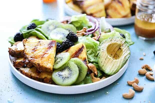 A white bowl on top of a blue surface filled with grilled pineapple wedges, kiwi slices, sliced avocado, thin slices of red onion, blackberries, salt greens, and cashews. There are cashews sprinkled on the blue surface next to a jar with dressing in it and a second white bowl of grilled pineapple wedges. 