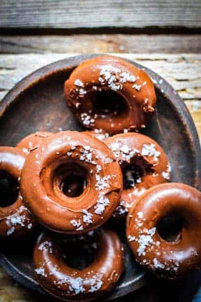 A dark brown wooden bowl full of chocolate peanut butter donuts that are iced with chocolate icing and sprinkled with flaky sea salt. The bowl is on top of a light gray wooden table