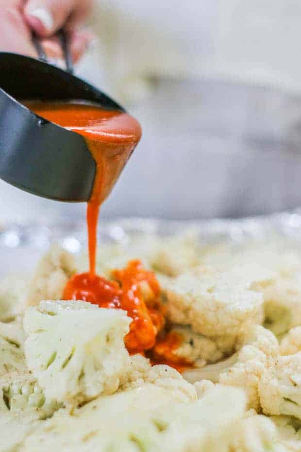 A woman in a cream colored shirt and tan linen apron pouring red buffalo sauce from a black measuring cup over a sheet pan of cauliflower florets.