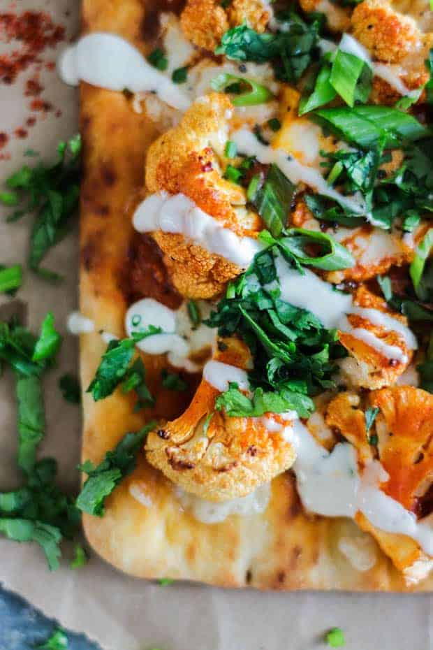 Close up of Roasted Buffalo Cauliflower Flatbread on top of a piece of brown butcher paper that has been layered over a metal table. The flatbread is piled high with orange buffalo cauliflower , drizzled with ranch dressing, and sprinkled with thinly sliced green onions and minced herbs.