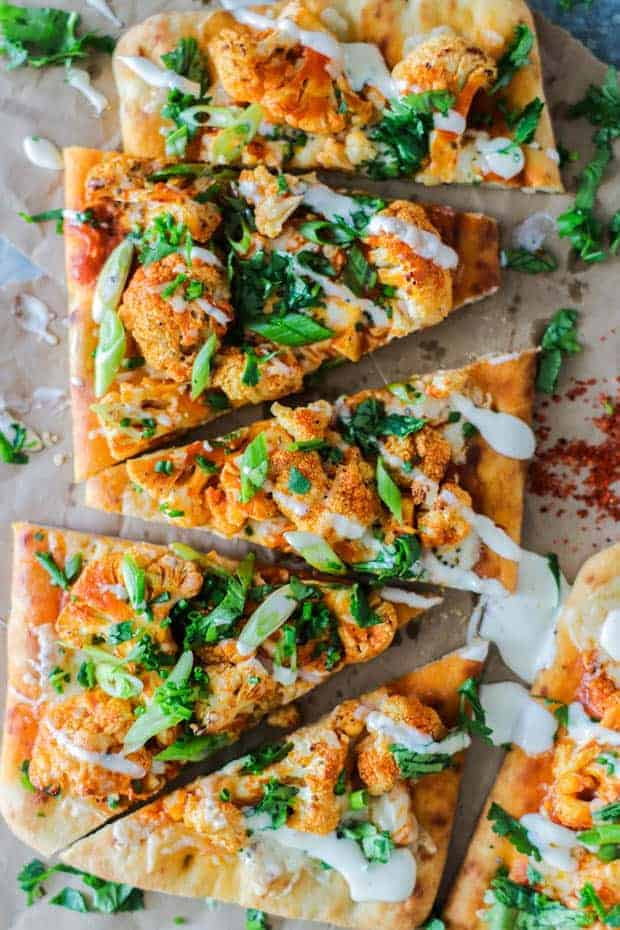 Triangles of Roasted Buffalo Cauliflower Flatbread on top of crinkled brown parchment paper. The flatbread is topped with orange buffalo flavored cauliflower, a drizzle of ranch dressing, green onion slices, and minced cilantro.