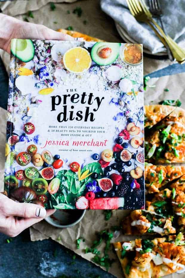 A woman hands holding a copy of The Pretty Dish cookbook over a metal table that is layered with brown parchment paper that has slices of Roasted Buffalo Cauliflower Flatbread on top.