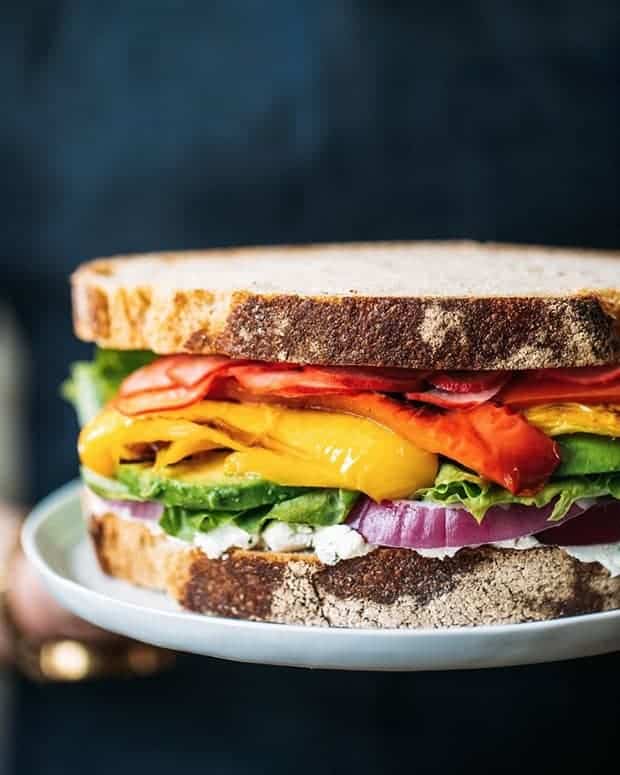 A person with a navy blue linen apron holding a white plate that has a very colorful sandwich on top. The sandwich is layered with a slice of bread, a white spread, red onions, greens, avocado, roasted yellow and red bell peppers, pickled radish slices, and another slice of bread. 