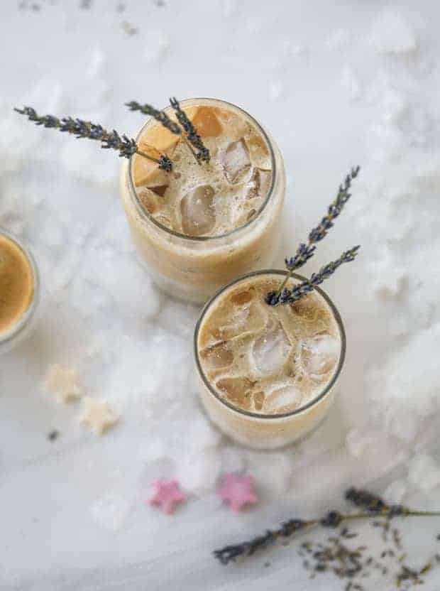 2 clear glasses filled with iced coffee on top of a marble surface. There are 2 lavender sprigs in each glass. There are lavender sprigs, ice cubes, and a few flower blossoms on the marble surface.