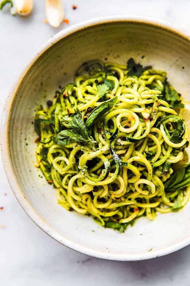 An off white bowl of zucchini "noodles" tossed in a green pesto. and a few basil leaves. The bowl sit son a white surface with a few garlic cloves on top.