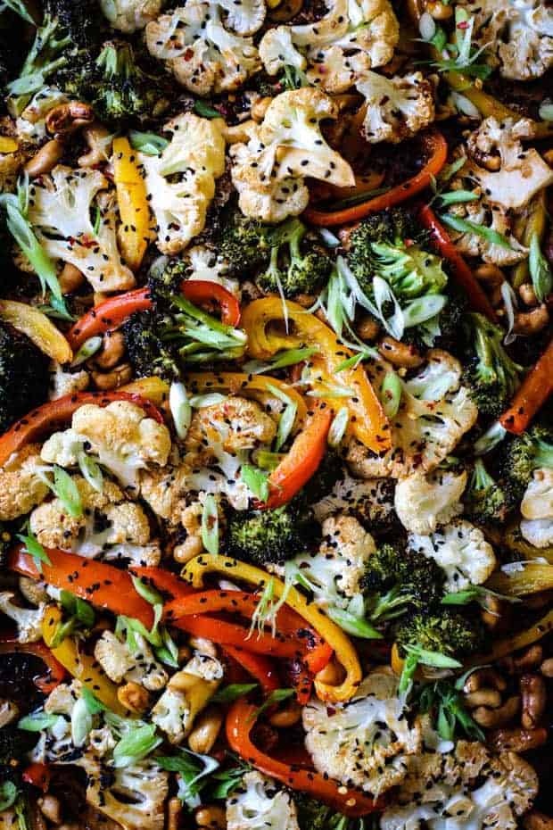 A close up of a sheet pan full of vegetables that have been roasted. There are cauliflower and broccoli florets, red and yellow bell pepper strips, and roasted cashews. Teh sheet pan is garnished with thin slices of green onion, black sesame seeds, chive blossoms, and basil blossoms.