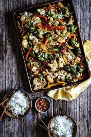 A large sheet pan filled with roasted vegetables like cauliflower, broccoli, red and yellow green peppers, and cashews sits on top of a yellow dish cloth on top of a wooden table. There are also 2 small brown bowls filled with white rice an da small bowl of red chili pepper flakes.