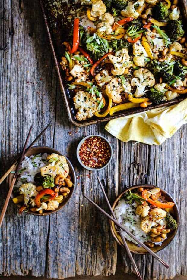 A sheet pan full of roasted cauliflower, broccoli, red and yellow green peppers, and cashews sits atop a yellow cloth napkin on a n old wooden table. There are 2 small brown bowls each filled with white rice and the roasted vegetables. Both bowls have a set of wooden chopsticks placed on them. There is a small dish of red pepper flakes in-between the two bowls of food.