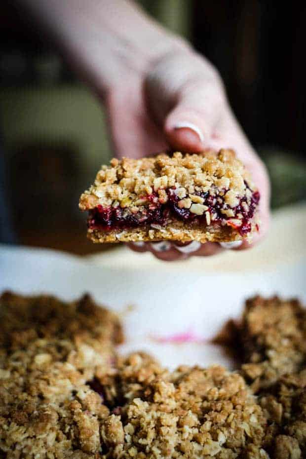 A woman's hand with white polished nails holding a cherry oat bar over a baking pan with more vbars in it. The bar has a golden crust, a vibrant red cherry filling, and a golden crumble oat crumble on top. 