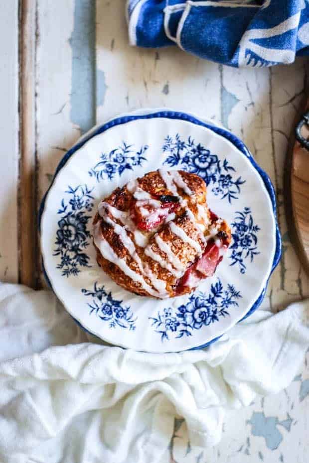 A white dessert plate with blue flowers painted around teh edges sits in the middle of an old painted table top. The table has white and light blue paint that is starting to chip and peel from the passage of time. There are 2 napkins on the table, one is white linen and one is blue with a subtle white design. On top of the dessert plate is a Strawberry and Cream Scone that is golden brown with roasted strawberries peeking out from the scone. There is a zig zag drizzle of white icing across the top of the scone.