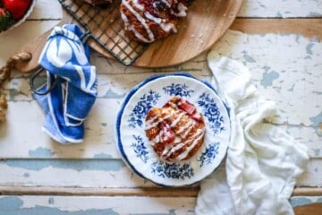 A white dessert plate with blue flowers painted around the edges sits in the middle of an old painted table top. The table has white and light blue paint that is starting to chip and peel from the passage of time. There are 2 napkins on the table, one is white linen and one is blue with a subtle white design. On top of the dessert plate is a Strawberry and Cream Scone that is golden brown with roasted strawberries peeking out from the scone. There is a zig zag drizzle of white icing across the top of the scone. In the corner of the image there is a round wooden cutting board that has a wire cooling rack on top with more scones.