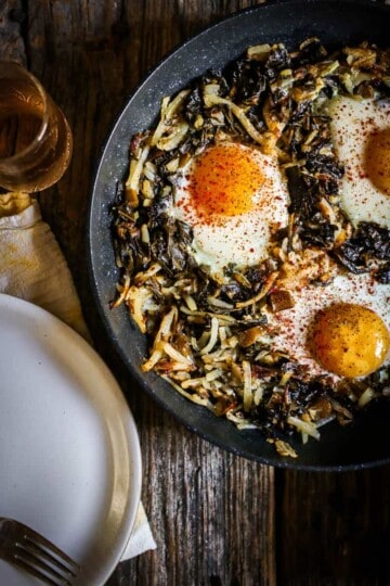 A skillet sits on top of weathered wooden table top. It is filled with a collard green and shredded potato hash and has 3 perfectly cooked eggs nestled inside the greens. There is also a white plate and a yellow napkin in the corner of teh image and a small glass of juice.