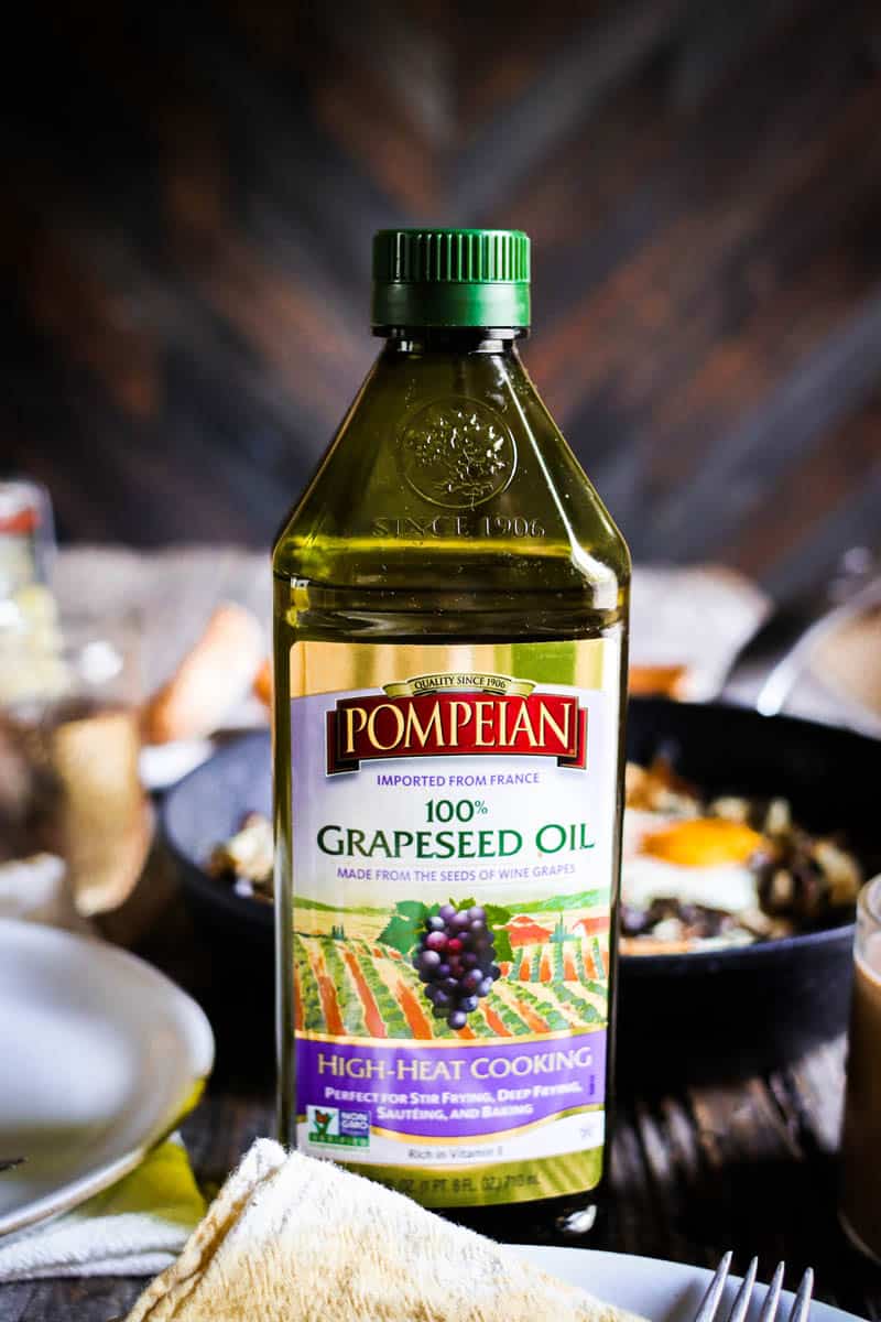 A bottle of Pompeian Garpeseed oil sits on a table top. There is a skillet filled with eggs and a collard green and shredded potato hash in teh background. There is also a small white wooden board with ciabatta rolls and a small jar of broccoli stalk pickles in the background
