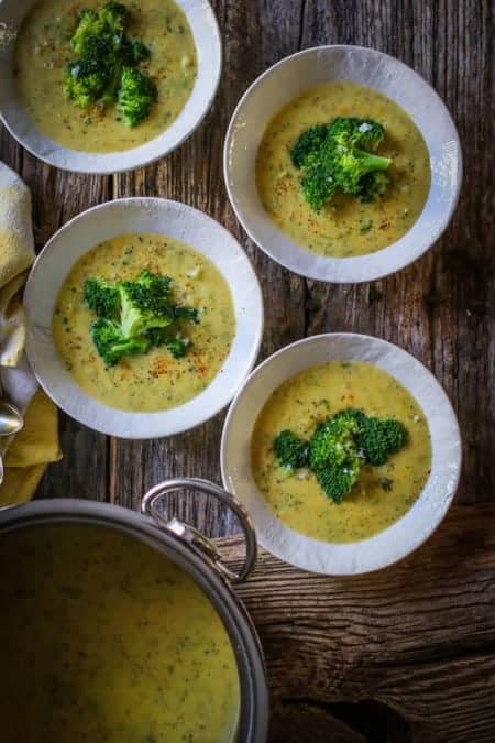 4 cream colored bowls are sitting on a weathered wooden table top . The bowls are filled with a golden colored broccoli cheddar soup. The soup is garnished with bright green blanched broccoli florets. There is also a large metal stock pot with teh soup in it and yellow and white cloth napkins on the table.