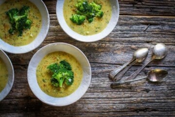 4 cream colored bowls are sitting on a weathered wooden table top . The bowls are filled with a golden colored broccoli cheddar soup. The soup is garnished with bright green blanched broccoli florets. There is a small pile of silver spoons on the table next to the bowls.