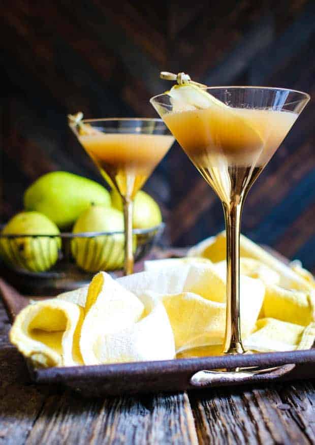 Two full martini glasses sit atop a rusted metal tray on top of a weathered wooden table top. The martini glasses are clear at the top and gold at the bottom of the glass and down the stem. Each martini has a slice of pear that has been bruleed floating in it. In the background there is a wire metal basket filled with green pears and in between the martini glasses is a yellow and white tea towel.