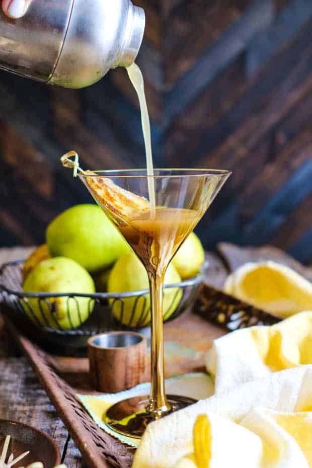 A rusted metal tray with a decorative edge sits atop a weathered wood table. There is a small wire metal basket in teh background that is filled with fresh green pears. In front of that is a martini glass that is clear at the top, but gold at the bottom of the glass and down the stem. There is a bruleed slice of pear laid inside the glass and someone is pouring the martini from the shaker into the glass. There are white and yellow cotton tea cloths in front of the martini glass.