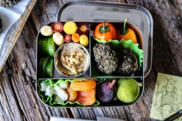 A stainless steel bento box packed with Mediterranean flavors of falafel, baby bell peppers, rainbow carrots, cucumbers, fresh figs, dried apricots.