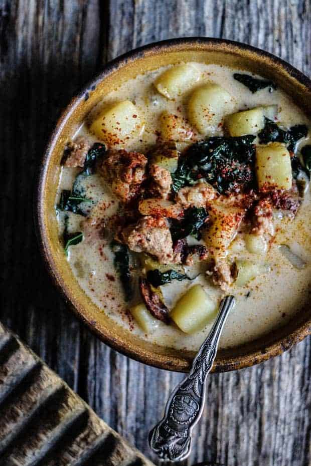 A light brown handmade soup bowl sits on a weathered wooden table top The bowl is filled with Zuppa Toscana soup, which is a creamy soup made from potatoes, sausage, and kale. There is an antique silver spoon in the bowl. The corner of a rustic chopping board is in the bottom left corner of the image.