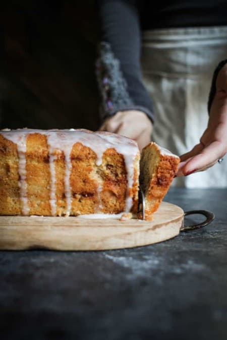 A golden brown loaf cake is on a wooden cutting board in top of a dark tabletop. The loaf cake has white icing drizzled on top and its dripping down the sides. A woman is standing in the background, she is wearing a tan linen apron and a black thermal shirt. The shirt has a decorative cuff around teh sleeve. The woman is slicing a piece of the loaf cake with a knife.