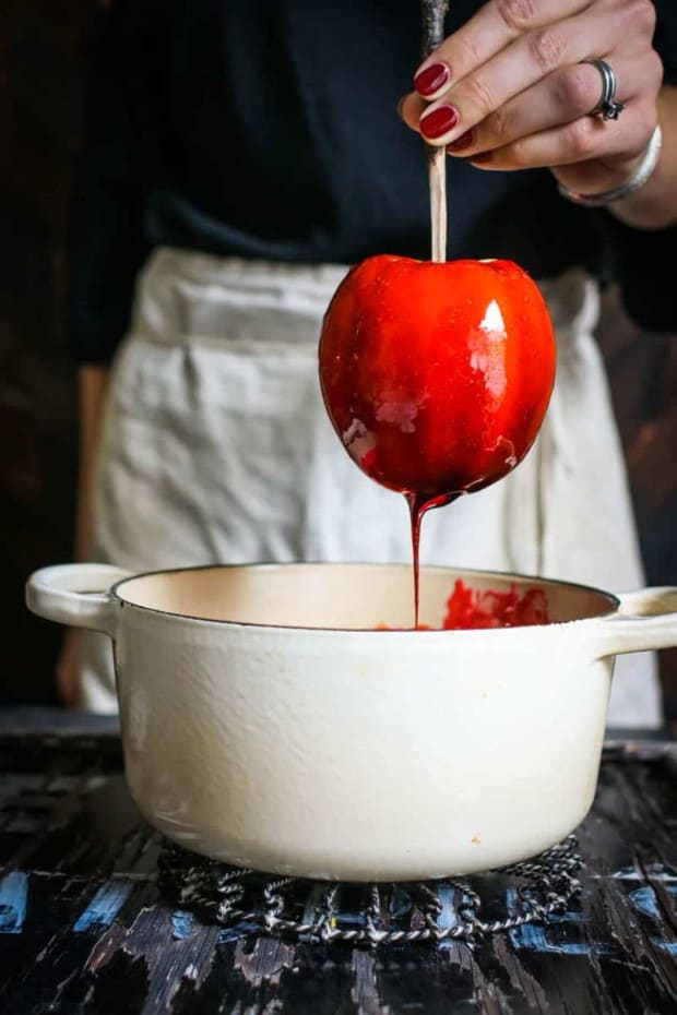 A woman wearing a black shirt and linen apron is dipping an apple on a stick into red candy syrup that is in an off white spot sitting on a black table top.