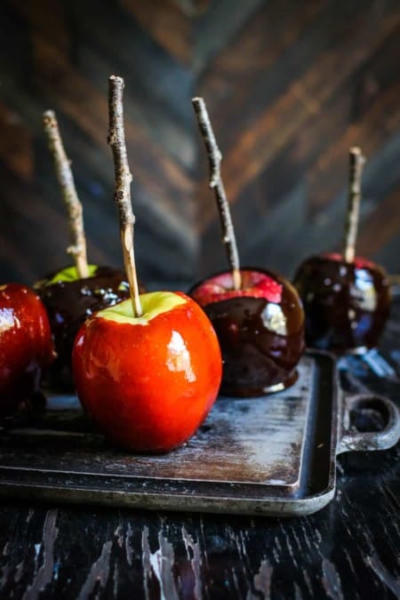 5 candy apples are sitting on top of a metal tray on a black table top. 2 of teh candy apples are red and 3 are black. The candy apple's sticks are made from real tree branches.