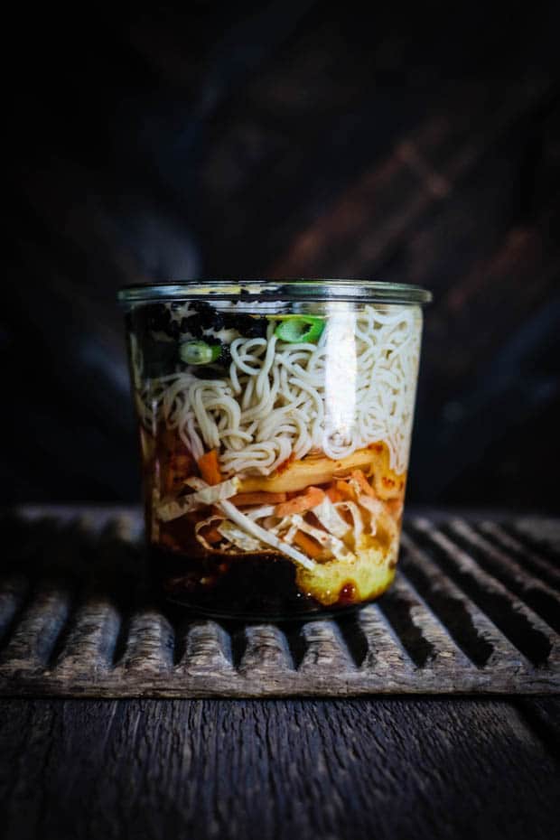 A glass week jar filled with the ingredients for messing gluten free cup of noodles with kimchi sits on a wooden table in front of a dark wood wall.