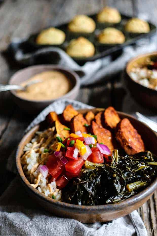 A bowl filled with brown rice, roasted sweet potatoes, collard greens, and pico de gallo sits on top of a line napkin on a table top. There is a small bowl of chipotle cashew cream in the background as well as pan of cornbread muffins. 