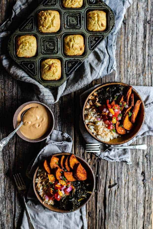 2 bowls filled with brown rice, collard greens, roasted sweet potatoes, and pic de gallo are on a wooden table top. There is a small bowl of chipotle cashew cream and a pan of cornbread muffins on testable as well as linen napkins and a few forks. 