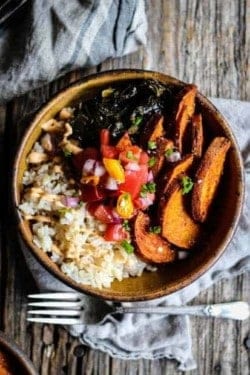 A handmade bowl filled with brown rice, sweet potatoes, collard greens, and pico de gallo is sitting on a wood table top with a fork and linen napkin next to it.