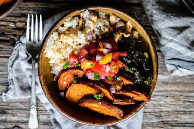 A handmade bowl filled with brown rice, sweet potatoes, collard greens, and pico de gallo is sitting on a wood table top with a fork and linen napkin next to it.